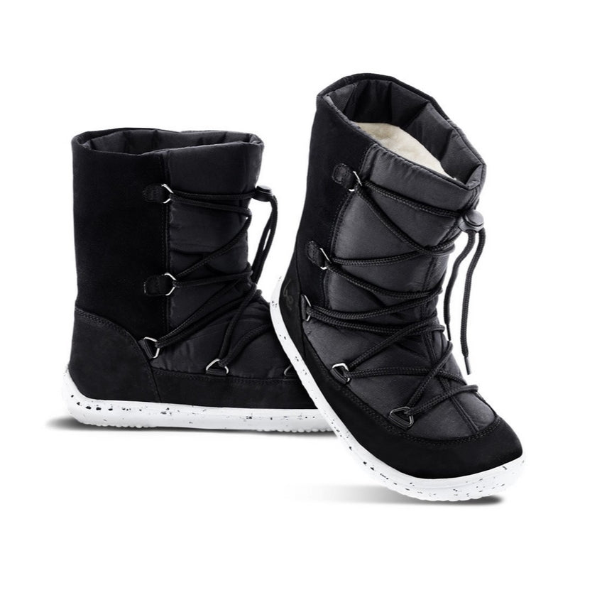 A photo of Be Lenka Kids Snowfox boots in black. Black suede and textile and light grey soles surround the outside. Black laces are widely connected from side to side starting at the ball of the foot and going until the top of the boot with a quick lace connector securing the laces in place. Left shoe is shown from the left side with the right shoe heel proped up on the midfoot of the left shoe against a white background. #color_black