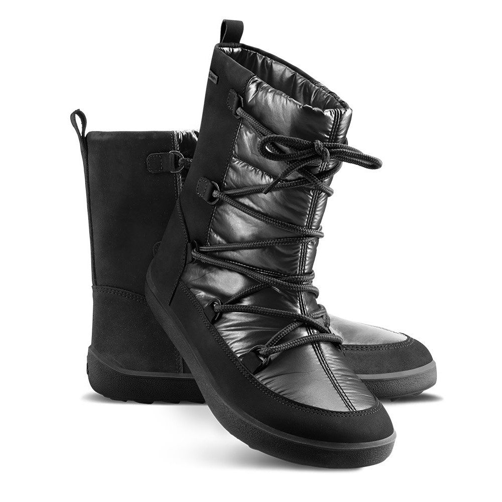 A photo of Be Lenka Adult Snowfox boots in all black. Black leather goes around the back with black satin over the top and front, and black soles. Laces are widely connected from side to side starting at the ball of the foot and going until the top of the boot. Left shoe is shown from the right side with the left boot propped up against it with a white background. #color_black