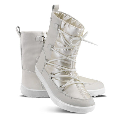 A photo of Be Lenka Adult Snowfox boots in Pearl White. Pearl white leather goes around the back with pearly white satin over the top and front, and white soles. Laces are widely connected from side to side starting at the ball of the foot and going until the top of the boot. Left shoe is shown from the right side with the left boot propped up against it with a white background. #color_pearl-white