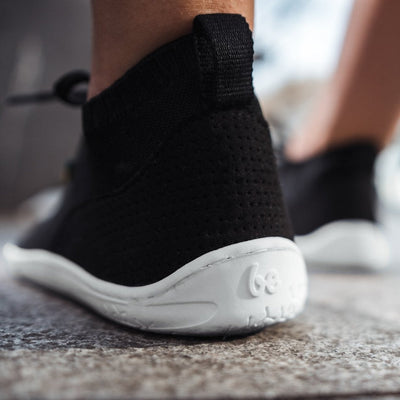A photo of black knit Be Lenka Stride Sneakers with white soles. A sock-like ankle opening is pulled on with a loop at the top of the opening. Soft perforated black microfiber accents the heel in a gentle hill shape. Laces are held on by a u-shaped microfiber sewn on top of the shoe. Left shoe is shown close up on the heel being worn by a man with faded black jogger jeans on top of a light grey gravel ground. #color_black