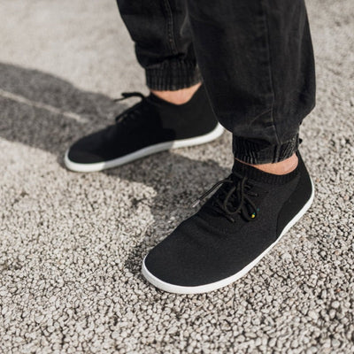 A photo of black knit Be Lenka Stride Sneakers with white soles. A sock-like ankle opening is pulled on with a loop at the top of the opening. Soft perforated black microfiber accents the heel in a gentle hill shape. Laces are held on by a u-shaped microfiber sewn on top of the shoe. Both shoes are shown being worn by a man with faded black jogger jeans diagonally from the left against a light grey gravel ground. #color_black