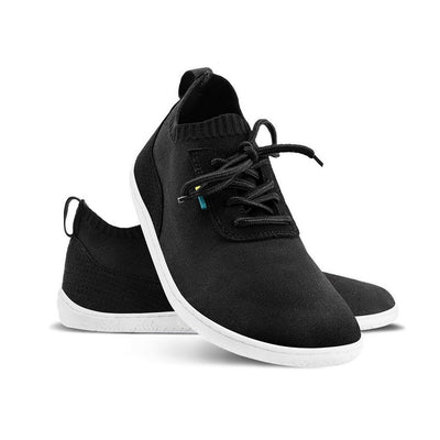 A photo of black knit Be Lenka Stride Sneakers with white soles. A sock-like ankle opening is pulled on with a loop at the top of the opening. Soft perforated black microfiber accents the heel in a gentle hill shape. Laces are held on by a u-shaped microfiber sewn on top of the shoe. Right shoe is shown propped up on the left shoe opening that is facing left against a white background. #color_black