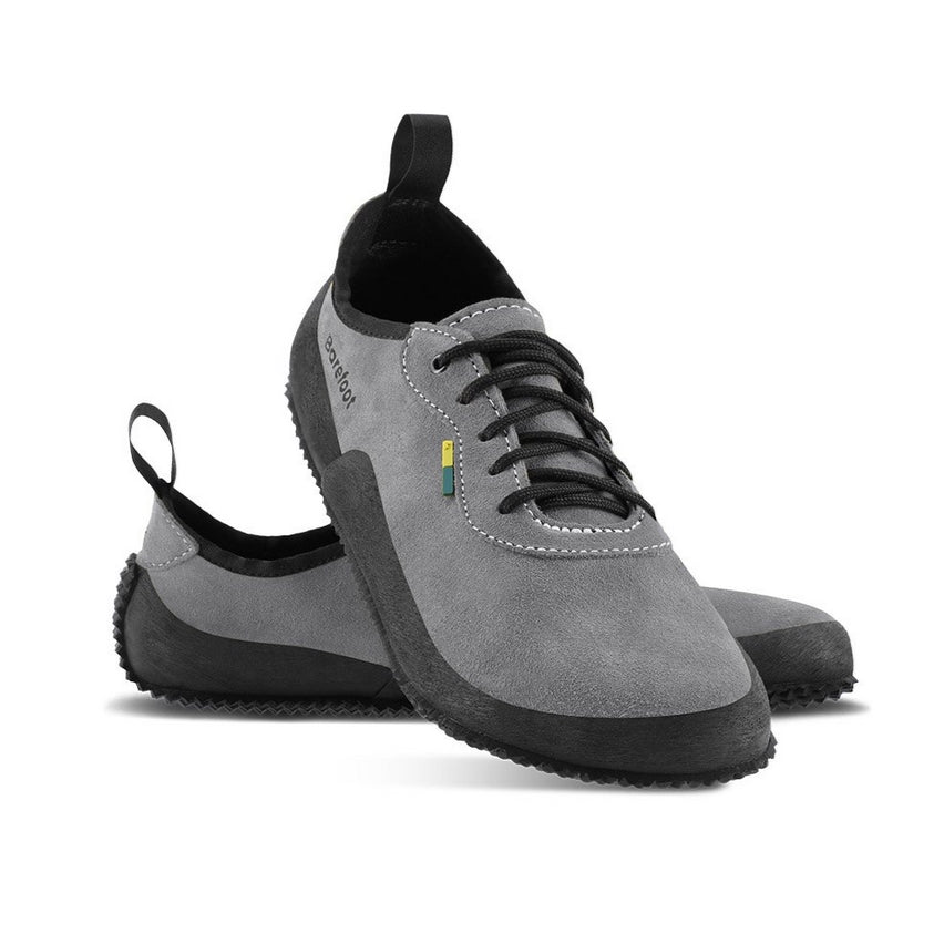 A photo of Be Lenka Trailwalkers 2.0 in Grey. Suede shoes are a low ankle height with black soles and laces, black rubber surrounding the lower part of the shoe, and pull/hook loops at the top of the heels. The top of the right shoe is shown tilted towards the camera here leaning on the left shoe pointing to the right against a white background. #color_grey