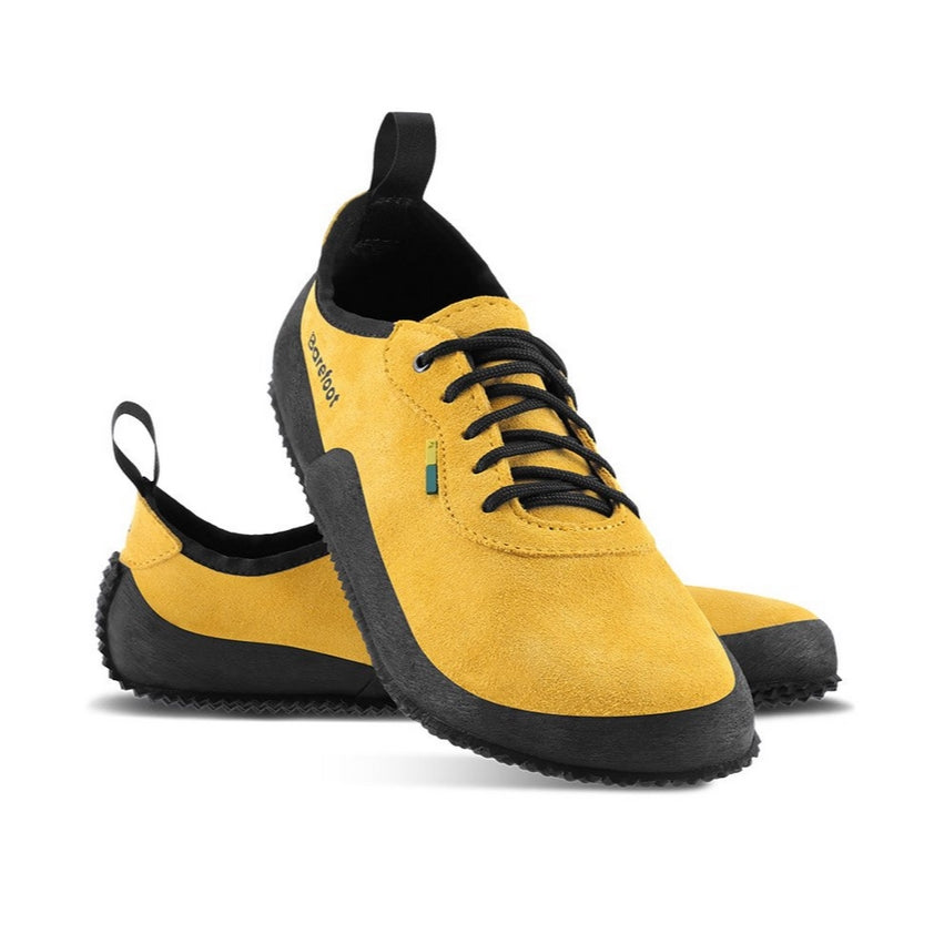 A photo of Be Lenka Trailwalkers 2.0 in Mustard. Suede shoes are a low ankle height with black soles and laces, black rubber surrounding the lower part of the shoe, and pull/hook loops at the top of the heels. The top of the right shoe is shown tilted towards the camera here leaning on the left shoe pointing to the right against a white background. #color_mustard