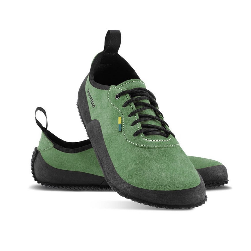 A photo of Be Lenka Trailwalkers 2.0 in Olive Green. Suede shoes are a low ankle height with black soles and laces, black rubber surrounding the lower part of the shoe, and pull/hook loops at the top of the heels. The top of the right shoe is shown tilted towards the camera here leaning on the left shoe pointing to the right against a white background. #color_olive-green