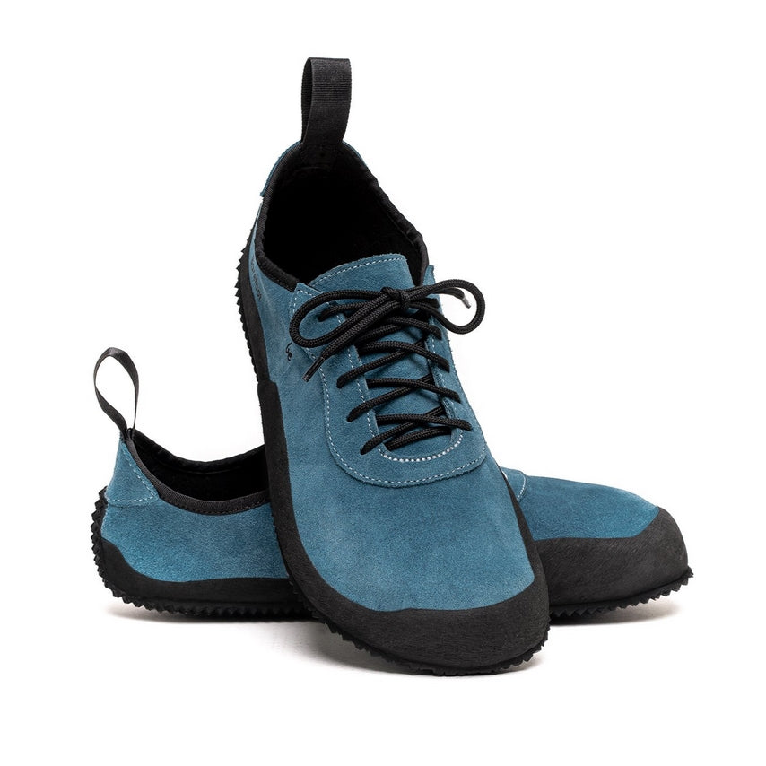 A photo of Be Lenka Trailwalkers in Deep Ocean blue. Suede shoes are a low ankle height with black soles and laces, black rubber surrounding the lower part of the shoe, and pull/hook loops at the top of the heels. The top of the right shoe is shown tilted towards the camera here leaning on the left shoe pointing to the right against a white background. #color_deep-ocean
