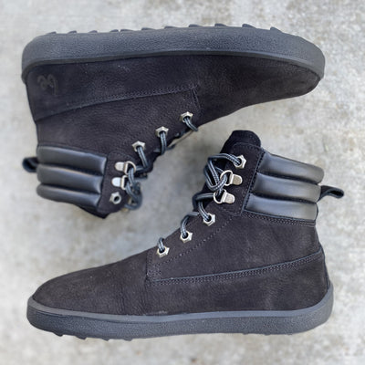 A photo of Be Lenka Nevada Boots made from nubuck leather and rubber soles. The boots are black in color, they are combat boot style with laces a padded color and a chunky eyelets. Both boots are shown laying on their sides opposite from each other against a concrete background. #color_black