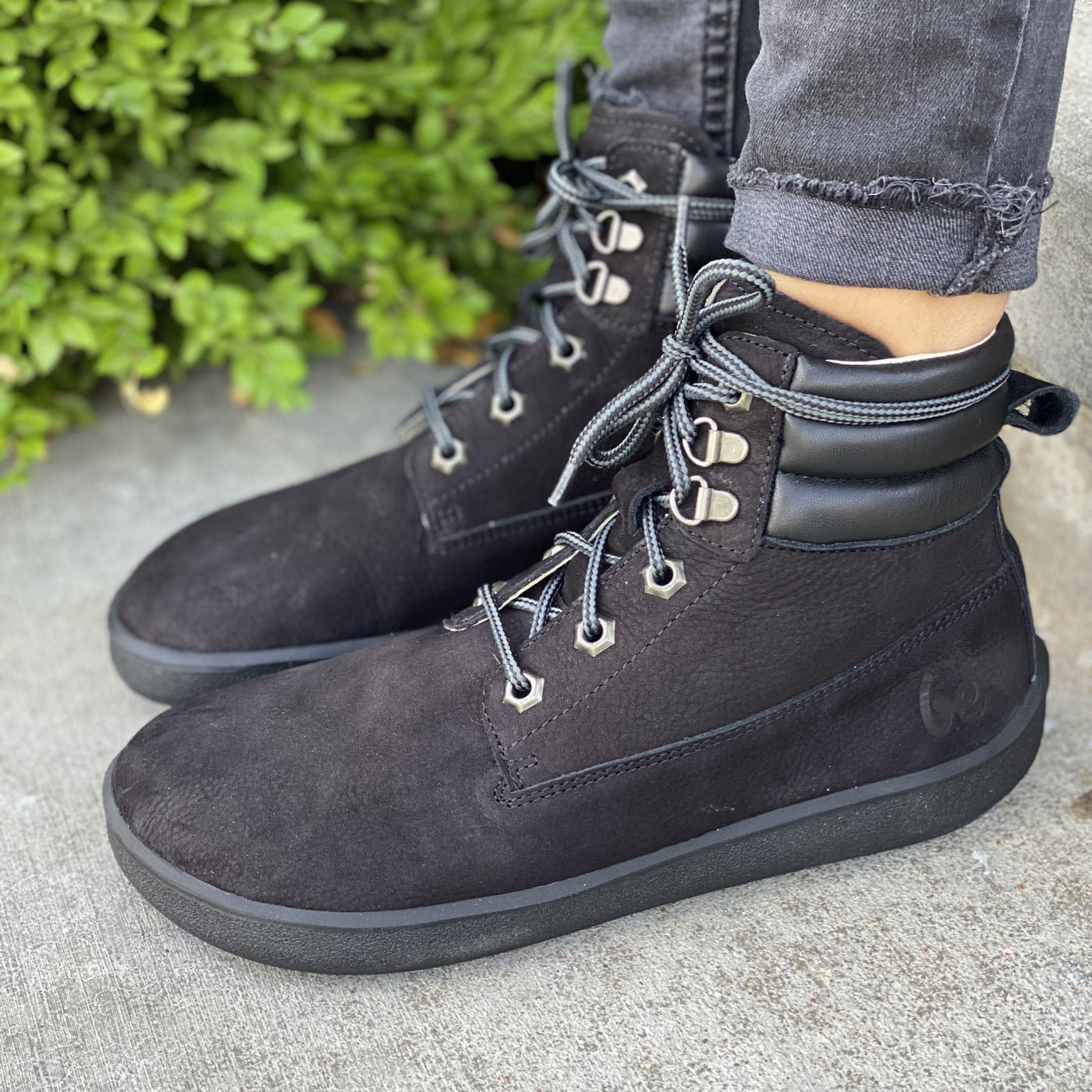 A photo of Be Lenka Nevada Boots made from nubuck leather and rubber soles. The boots are black in color, they are combat boot style with laces a padded color and a chunky eyelets. A woman is shown standing to the left on concrete from mid leg down wearing dark gray rolled jeans and the Nevada boots greenery is in the background. #color_black