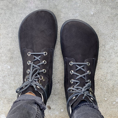 A photo of Be Lenka Nevada Boots made from nubuck leather and rubber soles. The boots are black in color, they are combat boot style with laces a padded color and a chunky eyelets. A woman is shown standing on concrete from above from mid leg down wearing dark gray rolled skinny jeans and the Nevada boots. #color_black