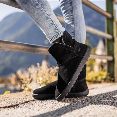 A photo of Belenka Polaris made from nubuck leather, sheepskin, and rubber soles. The boots are black in color they are a slip on with sheepskin inside. A woman is shown standing  from mid leg down wearing skinny jeans and the boots she is on pavement with a metal railing in front of her and a mountain is in the distance.  #color_black
