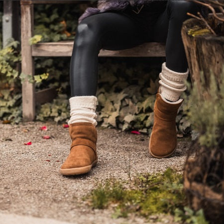A photo of Belenka Polaris made from nubuck leather, sheepskin, and rubber soles. The boots are brown in color they are a slip on with sheepskin inside. A woman is shown sitting on a bench she is shown from the knee down wearing black leggings, white socks, and the Polaris boots there is greenery in the background.  #color_brown