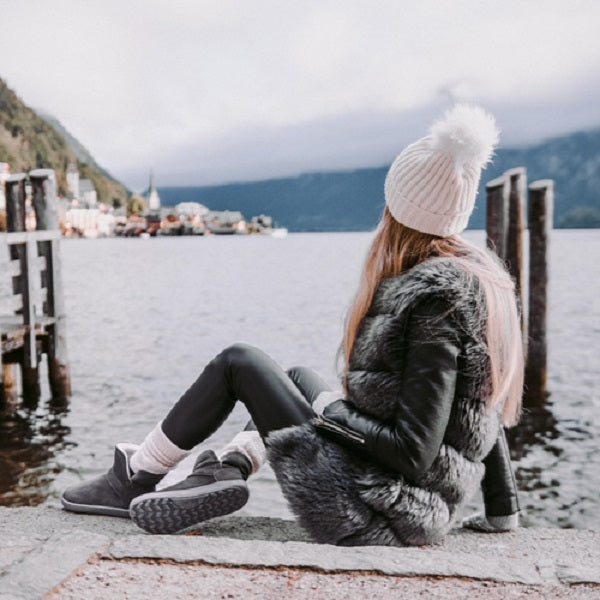 A photo of Belenka Polaris made from nubuck leather, sheepskin, and rubber soles. The boots are grey in color they are a slip on with sheepskin inside. A blonde woman is shown sitting on a dock in front of a lake, she is wearing a white beanie, a gray fuzzy vest, black shirt, black leggings, white socks, and the boots. She is shown looking in the distance at a town. #color_grey