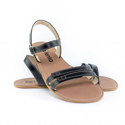 A photo of Black Be Lenka Summer Sandals made with leather and tan rubber soles. The sandals are black and have thin double straps on the front of the foot. They also have straps that go around the ankle and heel and have a small buckle. The right sandal is turned towards the front with its heel leaned on the left sandal against a white background in this photo. #color_black