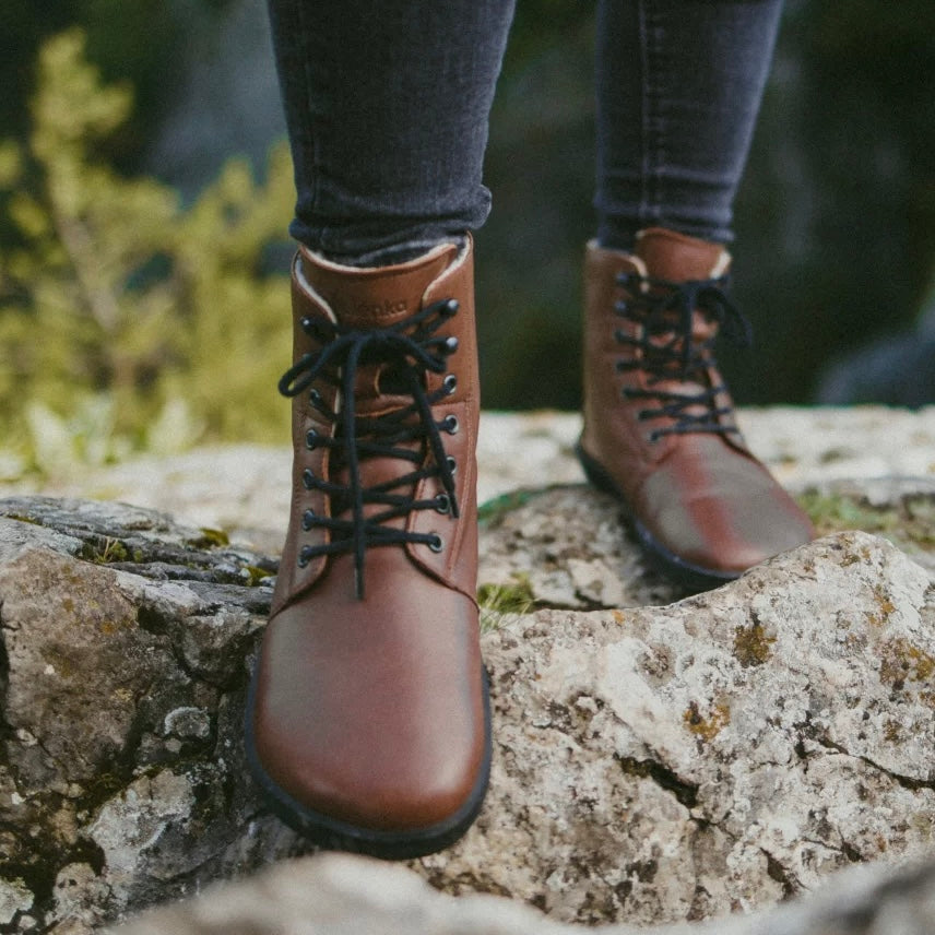 A photo of Be Lenka Winter Neo boots made with leather and rubber soles. The boots are brown in color and a lace up style with wool inside. A woman is shown from mid leg down wearing jeans and both Winter Neo boots. She is walking up hill on rocks. #color_brown-smooth-leather