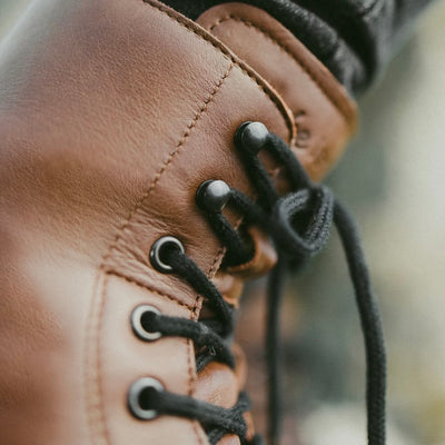 A photo of Be Lenka Winter Neo boots made with leather and rubber soles. The boots are brown in color and a lace up style with wool inside. One boot is shown up close by the laces and the background is blurry. #color_brown-smooth-leather