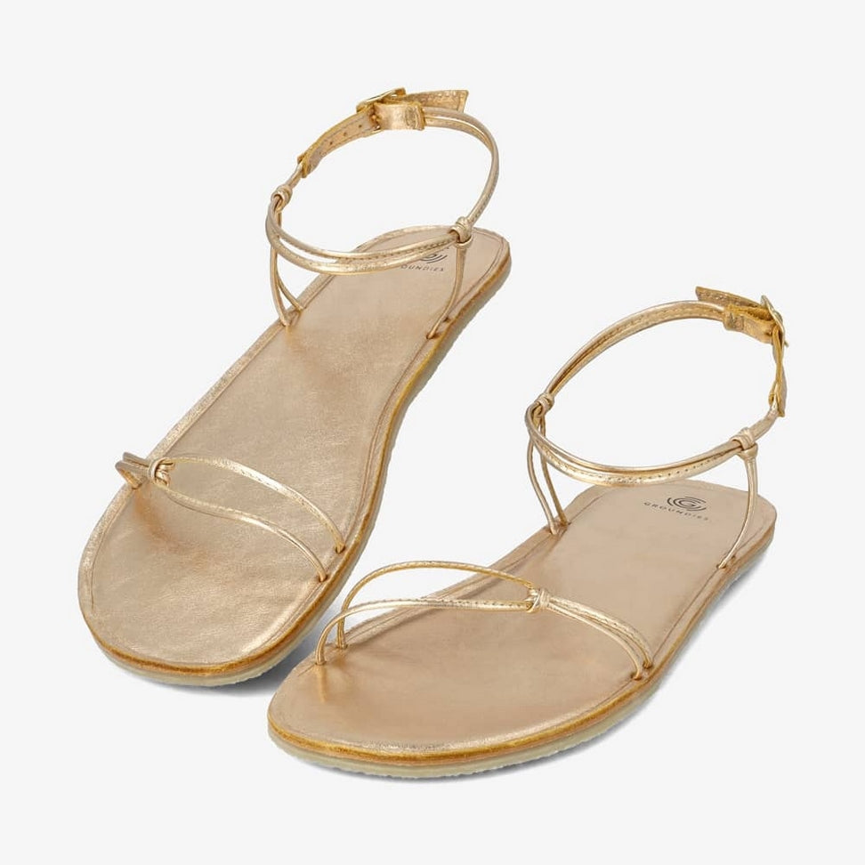 A photo of gold leather Groundies Cadiz Sandals. Two thin leather straps start at the ball of the big toe and end at the ball of the little toe with a twist detail by the second toe. An ankle strap made with two thin leather straps wraps around the ankle with a buckle closure attaching to the sole on either side of the foot. Shoes are shown diagonally from the left side against a white background. #color_gold