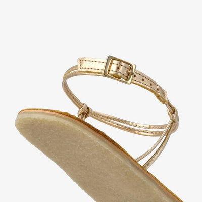 A photo of gold leather Groundies Cadiz Sandals. Two thin leather straps start at the ball of the big toe and end at the ball of the little toe with a twist detail by the second toe. An ankle strap made with two thin leather straps wraps around the ankle with a buckle closure attaching to the sole on either side of the foot. Shoe is shown floating diagonally from the back left heel as a close up on the sole and ankle strap against a white background. #color_gold