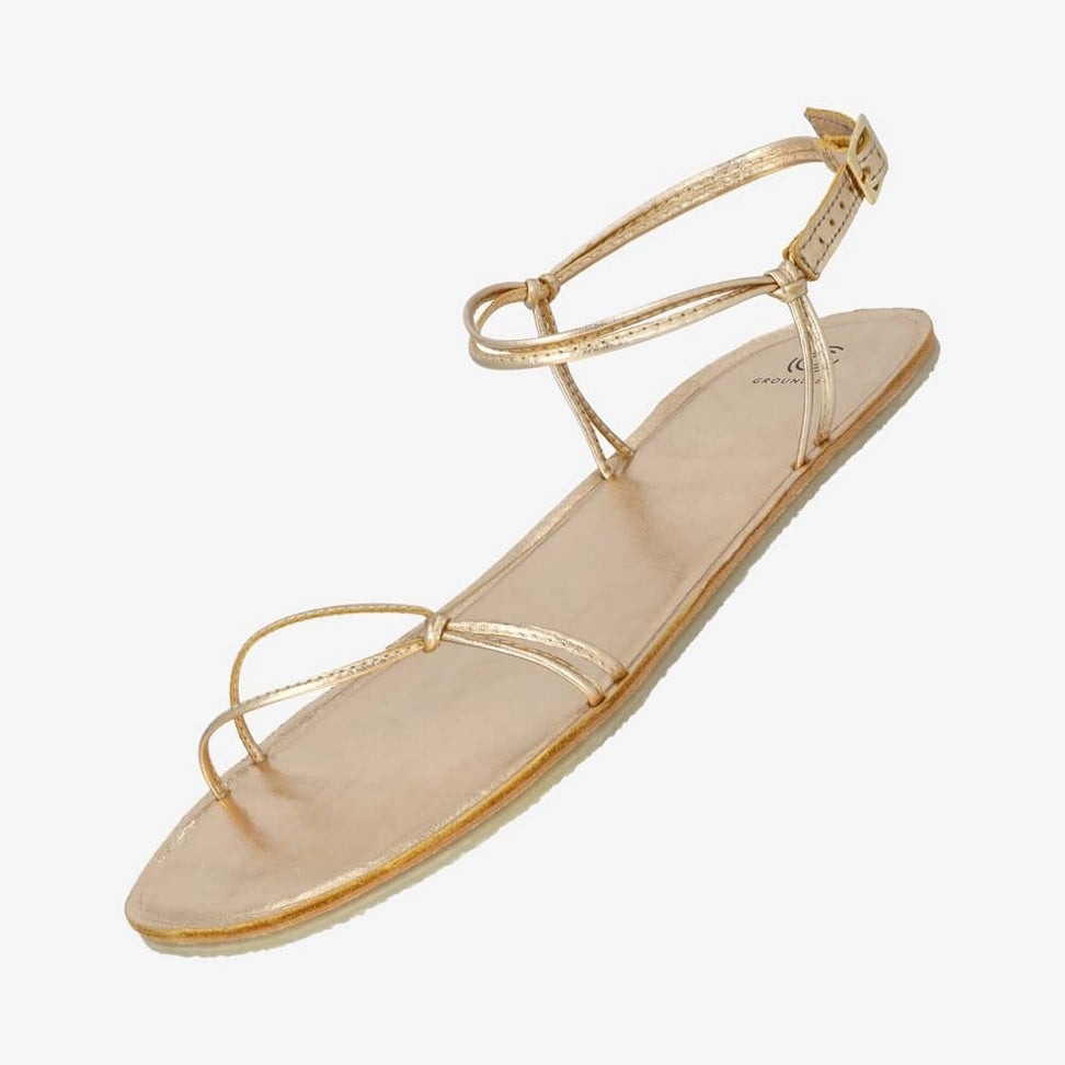 A photo of gold leather Groundies Cadiz Sandals. Two thin leather straps start at the ball of the big toe and end at the ball of the little toe with a twist detail by the second toe. An ankle strap made with two thin leather straps wraps around the ankle with a buckle closure attaching to the sole on either side of the foot. Shoe is shown floating diagonally from the left side against a white background. #color_gold