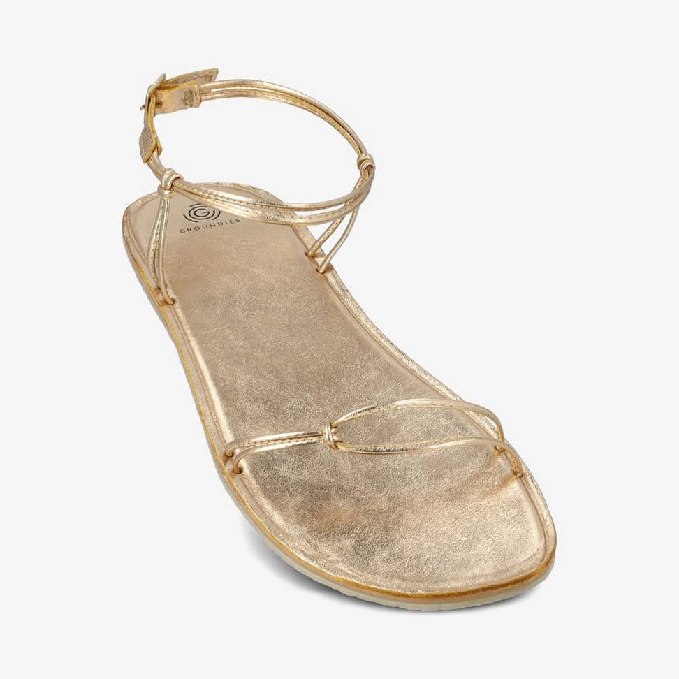 A photo of gold leather Groundies Cadiz Sandals. Two thin leather straps start at the ball of the big toe and end at the ball of the little toe with a twist detail by the second toe. An ankle strap made with two thin leather straps wraps around the ankle with a buckle closure attaching to the sole on either side of the foot. Shoe is shown tilted diagonally from the right side against a white background. #color_gold