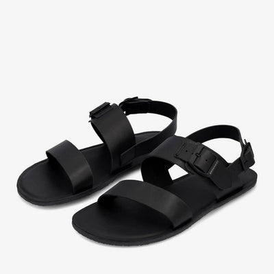 A photo of black leather Groundies Kos Sandals with thermo-rubber soles. One thick strap spans across the base of the toes. Another thick strap goes accross the arch of the foot with a big, black buckle closure. A thinner slingback strap keeps the sandals secure to the foot. Both shoes are shown diagonally from the left side against a white background. #color_black