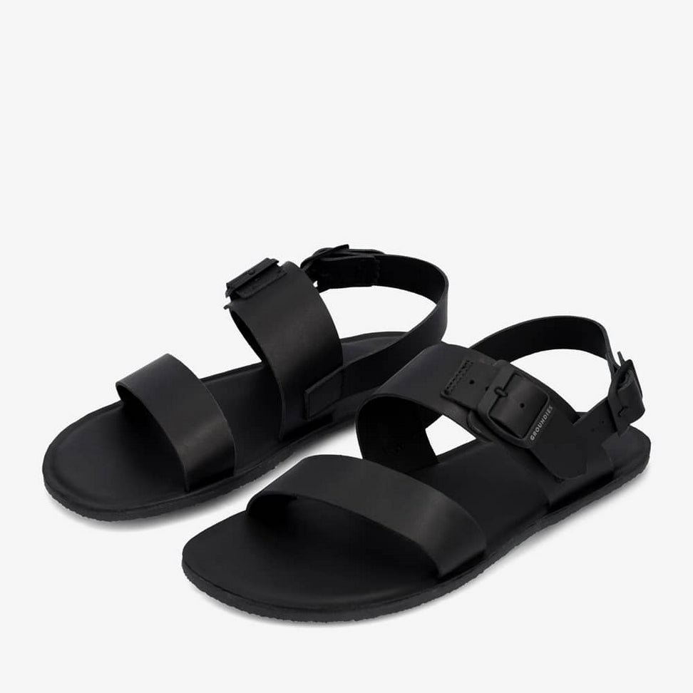 A photo of Groundies Kos thick strap sandals in black. Shoes have two thick leather straps - one around the base of the toes and another around the midfoot. A thiner black strap around the heel holds the shoe secure to the foot. The midfoot and ankle strap both have adjustable buckles. Both shoes are shown diagonally from the front left against a white background. #color_black