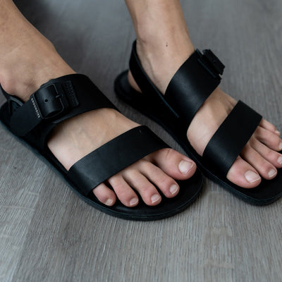 A photo of black leather Groundies Kos Sandals with thermo-rubber soles. One thick strap spans across the base of the toes. Another thick strap goes accross the arch of the foot with a big, black buckle closure. A thinner slingback strap keeps the sandals secure to the foot. Both shoes are shown on a womans foot diagonally from the front right sitting on a grey wooden floor. #color_black