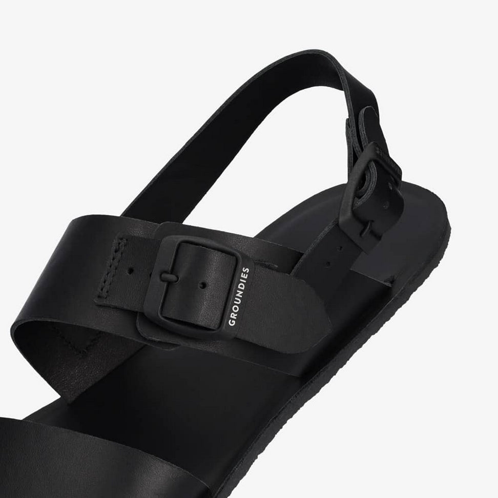 A photo of black leather Groundies Kos Sandals with thermo-rubber soles. One thick strap spans across the base of the toes. Another thick strap goes accross the arch of the foot with a big, black buckle closure. A thinner slingback strap keeps the sandals secure to the foot. Left shoe is shown floating from the diagonal left side close up on the buckle against a white background. #color_black