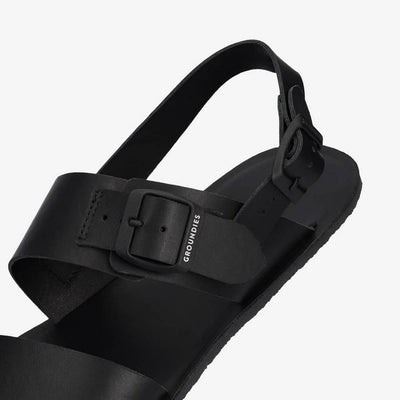 A photo of black leather Groundies Kos Sandals with thermo-rubber soles. One thick strap spans across the base of the toes. Another thick strap goes accross the arch of the foot with a big, black buckle closure. A thinner slingback strap keeps the sandals secure to the foot. Left shoe is shown floating from the diagonal left side close up on the buckle against a white background. #color_black