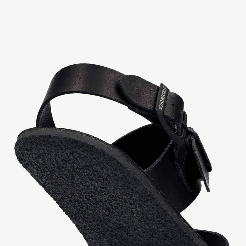 A photo of black leather Groundies Kos Sandals with thermo-rubber soles. One thick strap spans across the base of the toes. Another thick strap goes accross the arch of the foot with a big, black buckle closure. A thinner slingback strap keeps the sandals secure to the foot. Right shoe is shown floating from the diagonal right side close up on the slingback buckle and the sole against a white background. #color_black