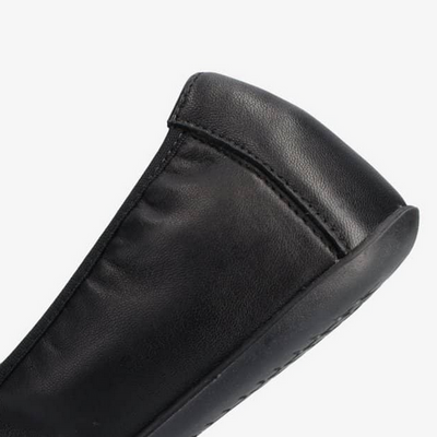 A photo of Groundies Lily Soft flats with a leather upper and black rubber true sense soles. The flats are a smooth leather in a black color with trim around the tops. The interior of the flats is a light beige color. The back left heel area of the flats are shown against a white background. #color_black