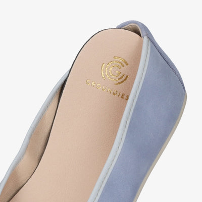 A photo of Groundies Lily Soft flats with a leather upper and cream rubber true sense soles. The flats are a nubuck in a light blue color, the trim around the tops of the flats is a baby blue color and is lighter than the rest of the flats. The interior of the flats is a light beige color. The right flat is shown with with a close up of the insole lining out of the shoe slightly to shown the detail against a white background. #color_light-blue