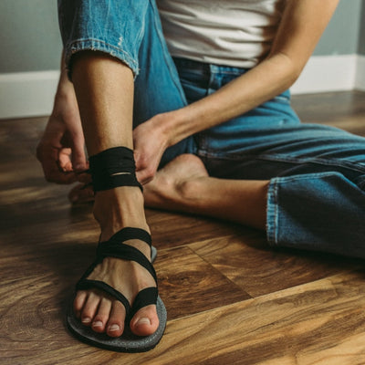 A photo of Juuri ribbon sandals made from leather, viscose, and rubber soles. The sandals have a grey footbed and long black laces. A woman is shown from waist down sitting on a wood floor wearing a white shirt, blue jeans, and one sandal that she is lacing up, a grey wall is in the background. #color_grey-black