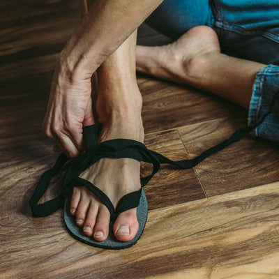 A photo of Juuri ribbon sandals made from leather, viscose, and rubber soles. The sandals have a grey footbed and long black laces. A woman is shown from mid leg down sitting on a wood floor wearing blue jeans putting on one of the sandals. #color_grey-black
