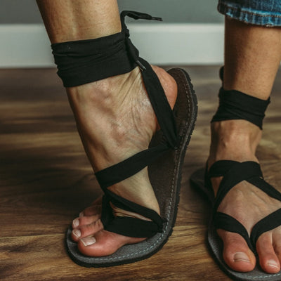 A photo of Juuri ribbon sandals made from leather, viscose, and rubber soles. The sandals have a grey footbed and long black laces. A woman is shown standing on a wood floor she is wearing blue jeans and both Juuri sandals, a grey wall is in the background. #color_grey-black