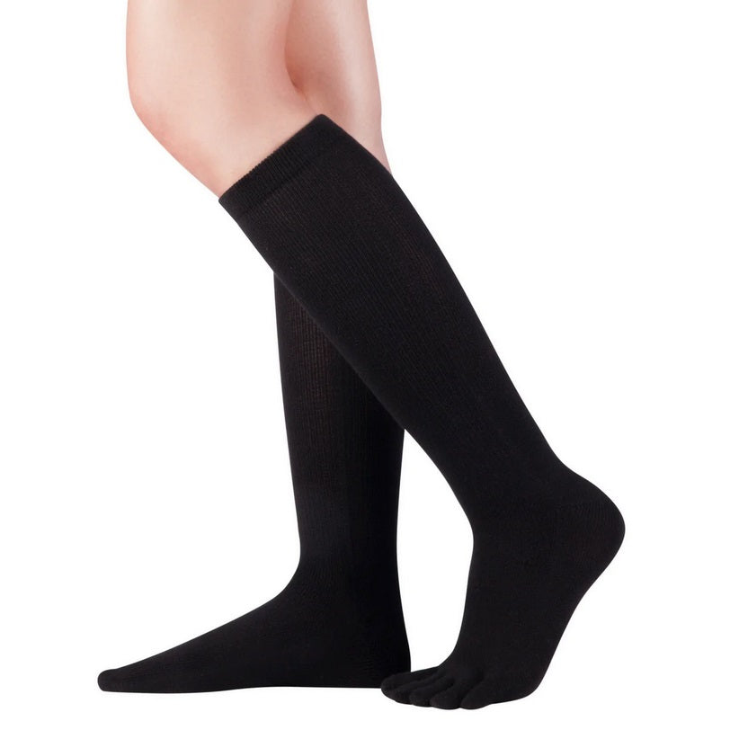A photo of Knitido knee high socks made with wool, cotton, polyester, nylon, and elastane. The socks are black in color. A woman is shown wearing the knee high socks standing to the right, she has the heel of one foot lifted and her leg slightly bent against a white background. #color_black