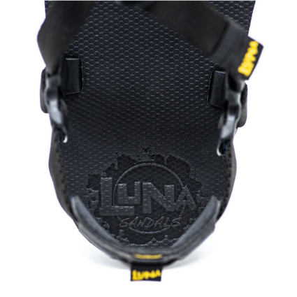 A photo of Luna Mono Winged Black Adventure Sandals made from a monkey grip technology footbed and a rubber sole. The sandals have a thong toe strap with straps that go around the ankle and heel. The sandals have a logo moon on the thong toe strap and luna logo on the heel and right ankle strap. The left sandal is shown up close from the back over the top showing the logo under the heel on the footbed against a white background. #color_black