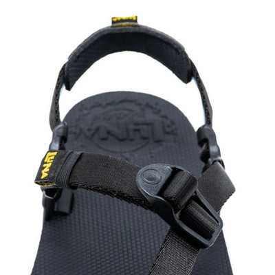 A photo of Luna Mono Winged Black Adventure Sandals made from a monkey grip technology footbed and a rubber sole. The sandals have a thong toe strap with straps that go around the ankle and heel. The sandals have a logo moon on the thong toe strap and luna logo on the heel and right ankle strap. The left sandal is shown up close angled from the back over the top showing the logo under the heel on the footbed against a white background. #color_black