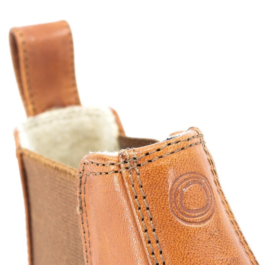 A photo of Mukishoes felted-wool-lined, leather Chelsea boots in caramel. Boots have brown elastic pannels on both sides of the shoe and a pull tab at the top of the ankle opening. Soles are a natural gum rubber color and are stitched to the leather. The right boot ankle opening is shown close up here from the front against a white background. #color_caramel