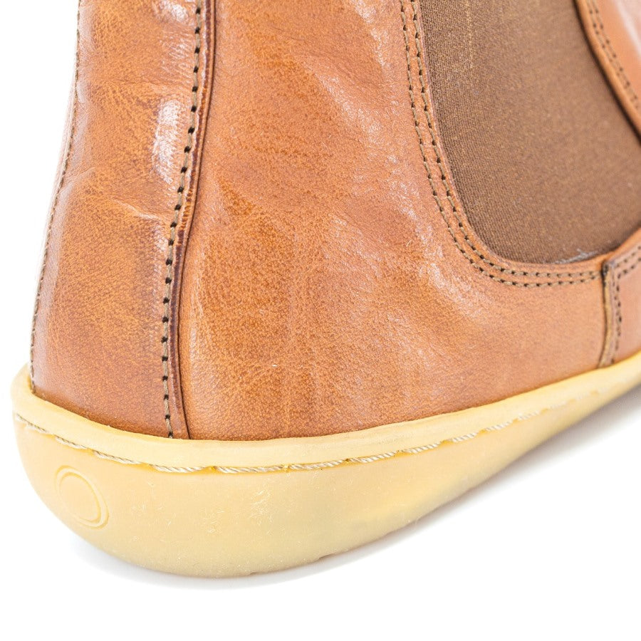 A photo of Mukishoes felted-wool-lined, leather Chelsea boots in caramel. Boots have brown elastic pannels on both sides of the shoe and a pull tab at the top of the ankle opening. Soles are a natural gum rubber color and are stitched to the leather. The right boot sole and heel is shown close up here diagonally from the right side against a white background. #color_caramel