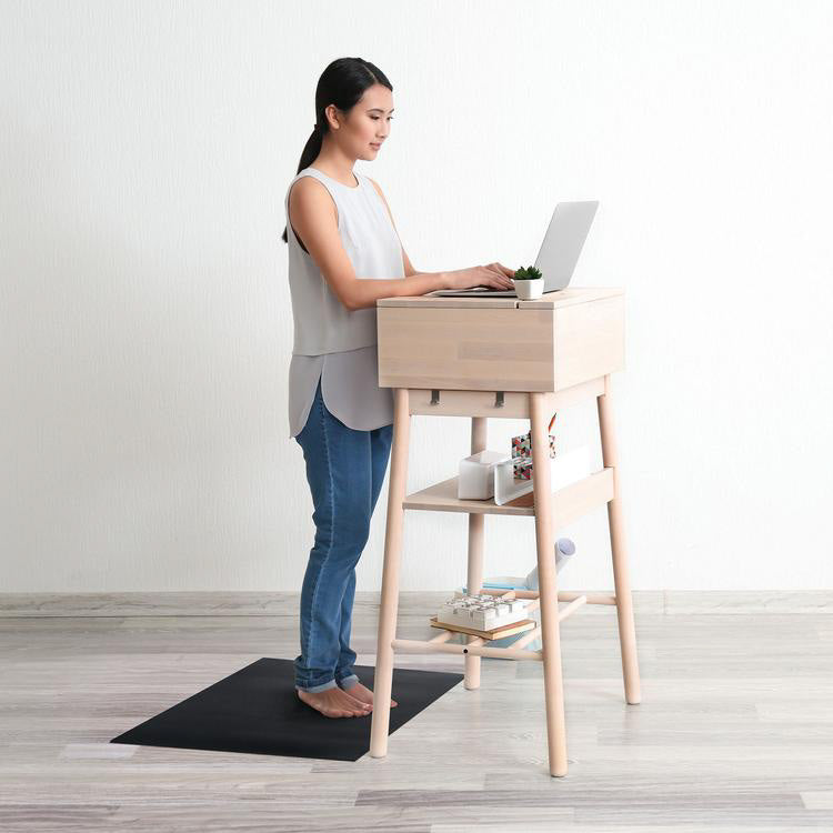 A photo of the Naboso standing mat a textured mat. The mat is black in color and has Naboso written on it. A dark haired woman is shown standing typing at a standing desk, she is wearing a white tank top and jeans while standing on the mat in a neutral colored room.