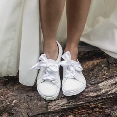 A photo of white Peerko Celebrate sneakers made with leather and recyclable soles. Sneakers have a slight sparkle and laces are ribbon. Shown from the front being worn by a woman in a wedding dress standing on a log. #color_white