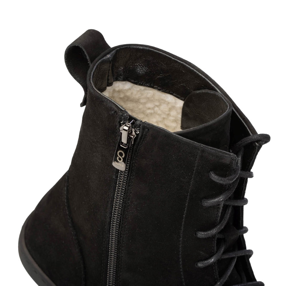 A photo of the Shapen Cozy lace up boots made from a water resistant nubuck leather upper, wool lining, and black rubber soles. The boots are black in color with black laces and have a zipper on the inner side. The left boot is shown from the top focused on the opening of the boot to show the wool lining and the zipper up close, on a white background. #color_black