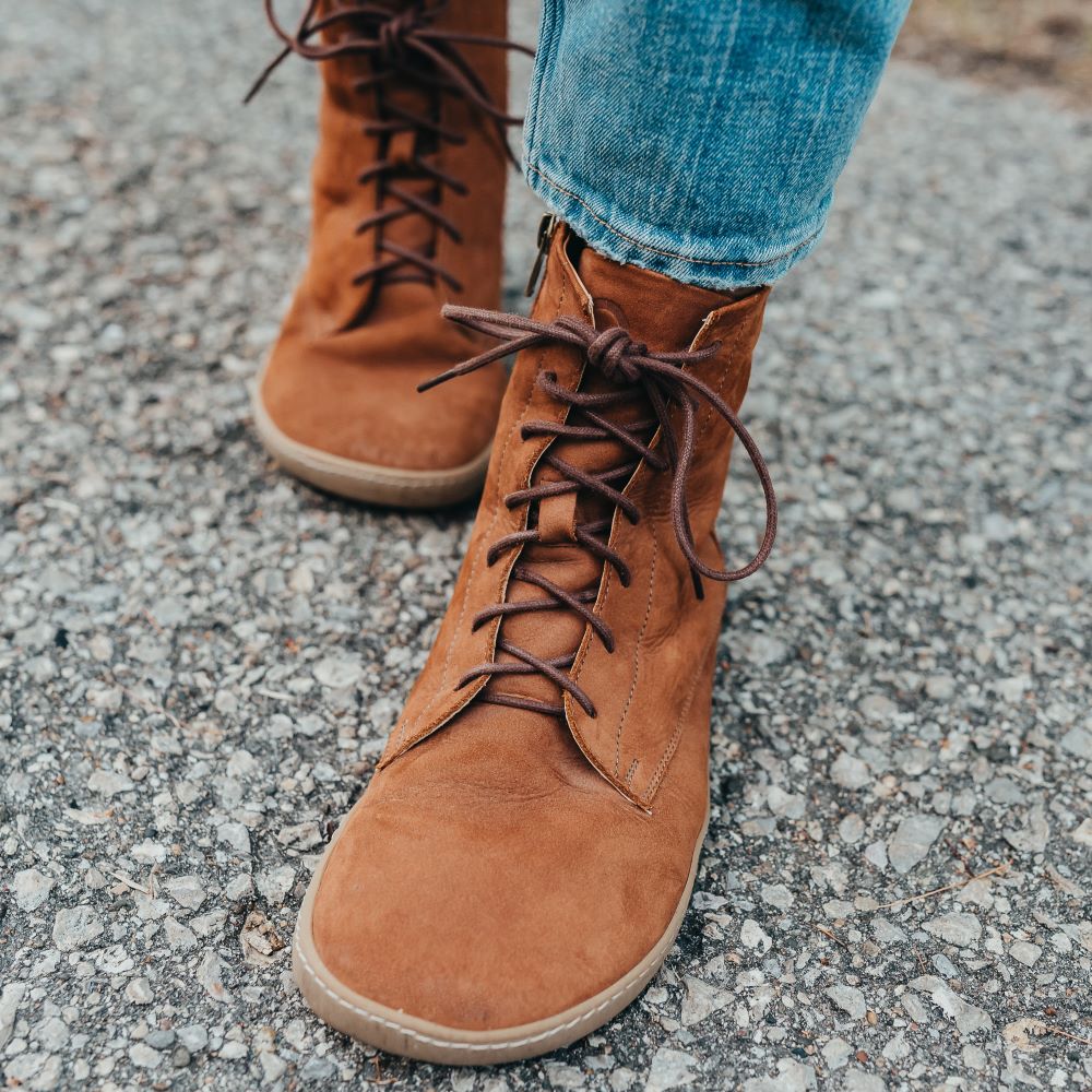 A photo of the Shapen Cozy lace up boots made from a water resistant nubuck leather upper, wool lining, and brown rubber soles. The boots are brown in color with brown laces and have a zipper on the inner side. Both boots are shown from the front on a woman's feet with a view of her ankles down. The woman is wearing blue skinny jeans and is walking on an asphalt road. #color_brown