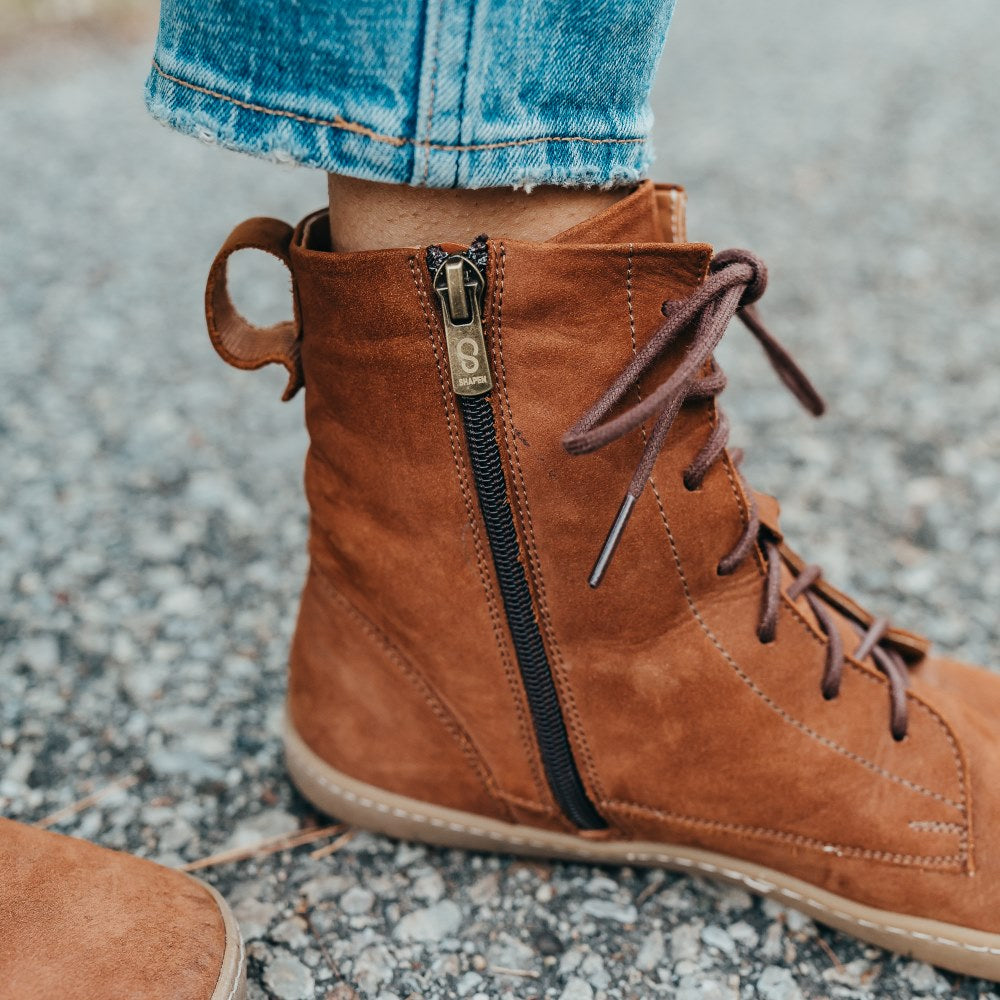 A photo of the Shapen Cozy lace up boots made from a water resistant nubuck leather upper, wool lining, and brown rubber soles. The boots are brown in color with brown laces and have a zipper on the inner side. The left boot is shown from the left side on a woman's feet with a view of her ankles down, showing the zipper detail. The woman is wearing blue skinny jeans and is standing on an asphalt road. #color_brown