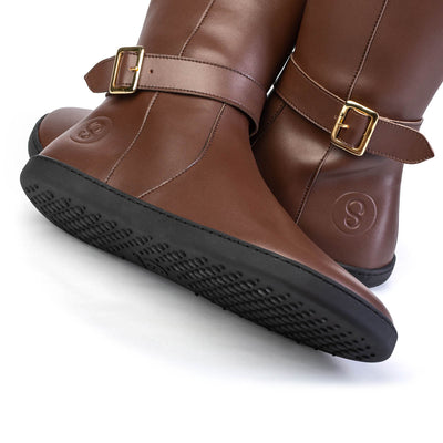 A photo of Shapen Glam lined riding boots made with vegan leather a faux fur lining and rubber soles. The boots are brown in color with gold buckles on the top and around the ankle, the ankle buckle strap is removable. Both boots are shown up close beside each other the one boot is laying over on top of the other facing the opposite direction against a white background. #color_brown