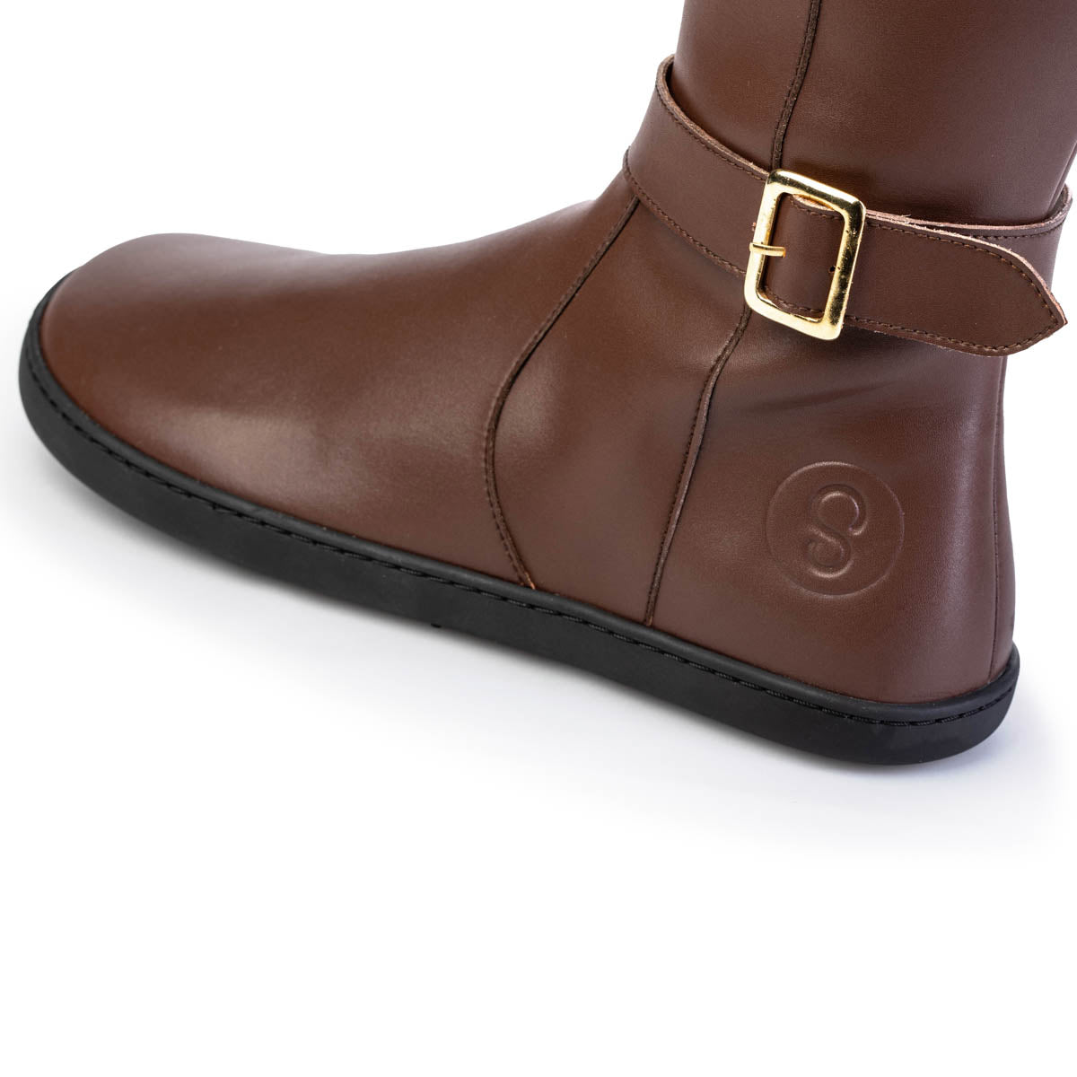 A photo of Shapen Glam lined riding boots made with leather a faux fur lining and rubber soles. The boots are brown in color with gold buckles on the top and around the ankle, the ankle buckle strap is removable. The left shoe is shown up close from the left side against a white background. #color_brown