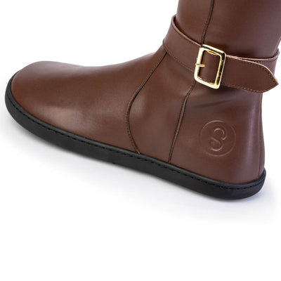 A photo of Shapen Glam lined riding boots made with vegan leather a faux fur lining and rubber soles. The boots are brown in color with gold buckles on the top and around the ankle, the ankle buckle strap is removable. The left shoe is shown up close from the left side against a white background. #color_brown