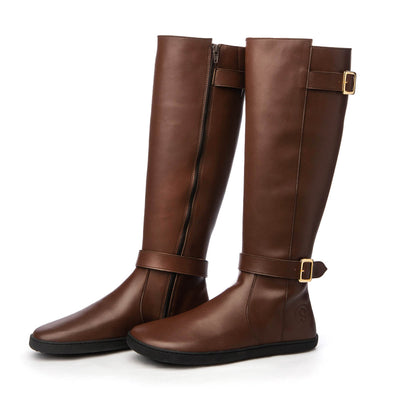 A photo of Shapen Glam lined riding boots made with leather a faux fur lining and rubber soles. The boots are brown in color with zippers on the side and gold buckles on the top and around the ankle, the ankle buckle strap is removable. Both boots are shown from the right side beside each other angled slightly towards the front against a white background. #color_brown