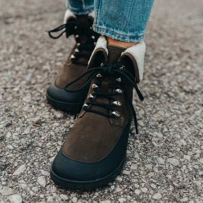 A photo of the Shapen Lynx lace up boots made from a water-resistant nubuck leather uppers and black rubber soles. The boots are dark green in color, have a black leather rim around the bottom inch of the shoe, black laces, and a white wool lining that can be shown by folding over the top of the boots. Both boots are shown  from the front on a woman's feet with a view of her shins down. The woman is wearing blue skinny jeans and is standing on an asphalt road. #color_dark-green 