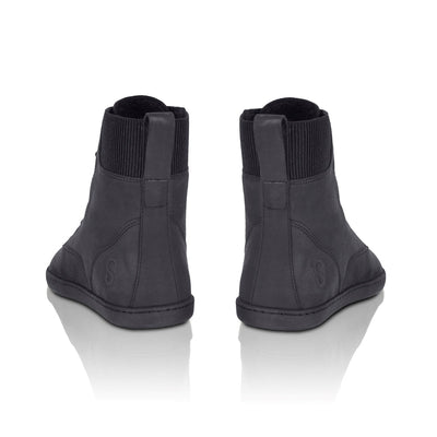 A photo of the Shapen Urbaneer combat boots made with nubuck leather, a thin wool lining, and rubber soles. The boots are black in color and have an elasticized panel on the top back of the boot. Both boots are shown from behind to show the elasticized panel, on a white background. #color_black